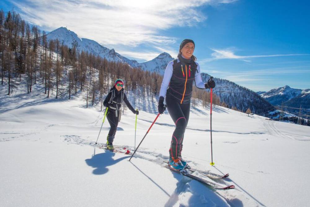Two skiers navigate a snowy landscape with tall mountains and a bright blue sky in the background, surrounded by trees.