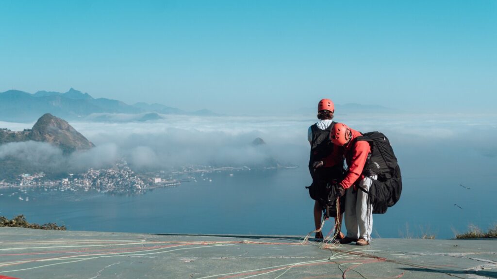 Two paragliders prepare their gear atop a mountain overlooking a misty lake and distant mountains under a clear blue sky.