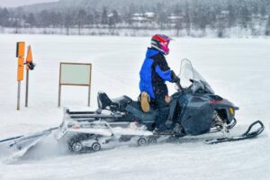 A person rides a snowmobile across a snowy landscape, with caution signs and a blank signpost visible in the background.