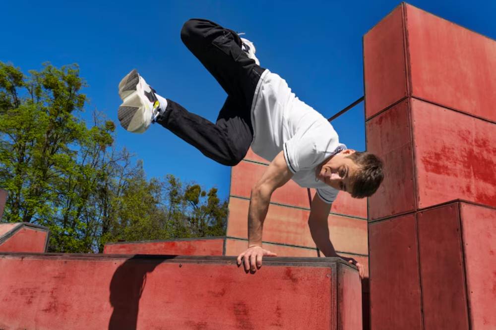 A person performs a dynamic parkour move, leaping between large red structures against a clear blue sky.