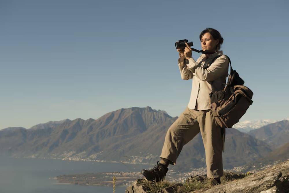 A person stands on a rocky ledge, holding a camera, with a panoramic view of mountains and a lake in the background.