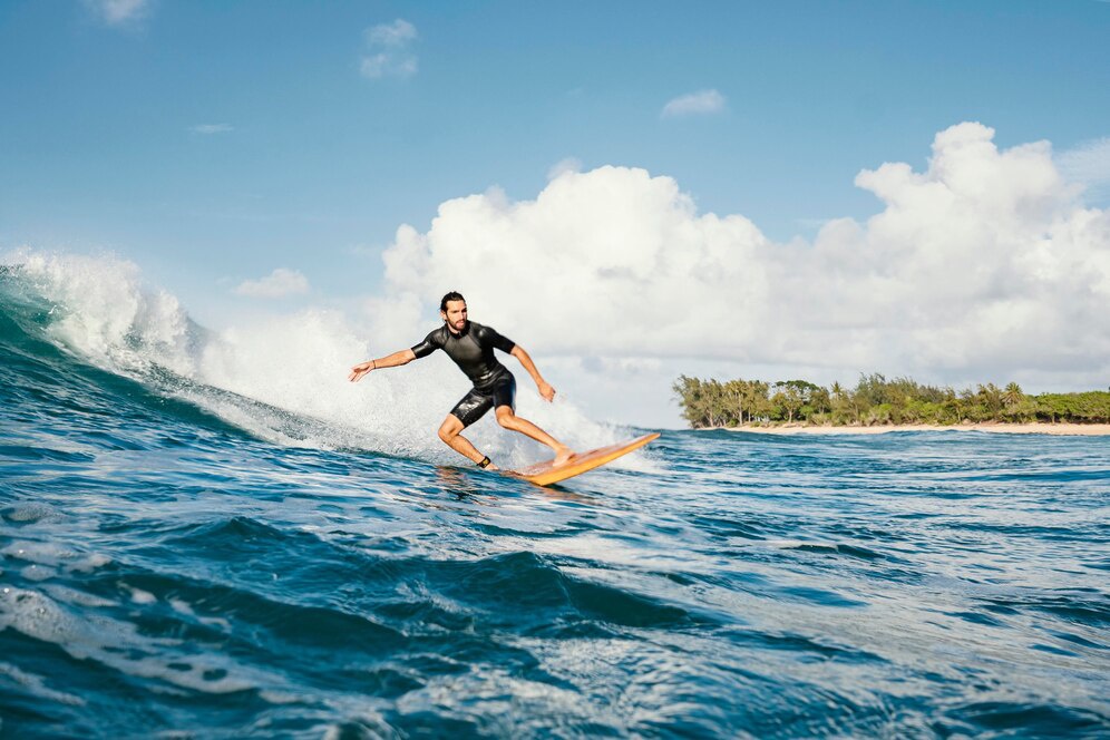 A surfer skillfully rides a wave on a sunny day, with lush greenery and clouds visible in the background.