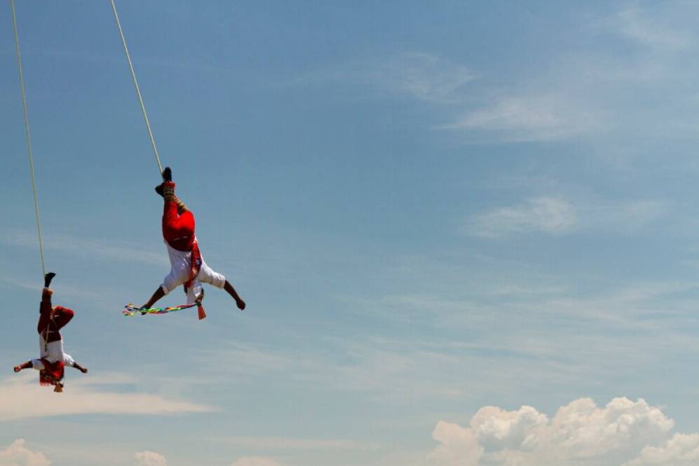 Two performers in traditional attire dangle from ropes, gracefully twisting against a bright blue sky with fluffy clouds.