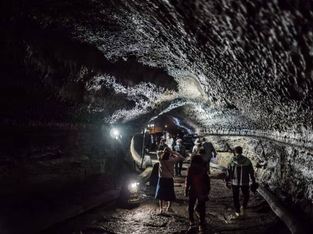 Tourists explore a dimly lit volcanic cave, with rough, textured walls and glowing lamps illuminating the rocky path ahead.