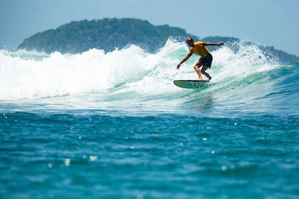 A surfer in a yellow shirt skillfully rides a wave, with a lush green island in the background under a clear blue sky.