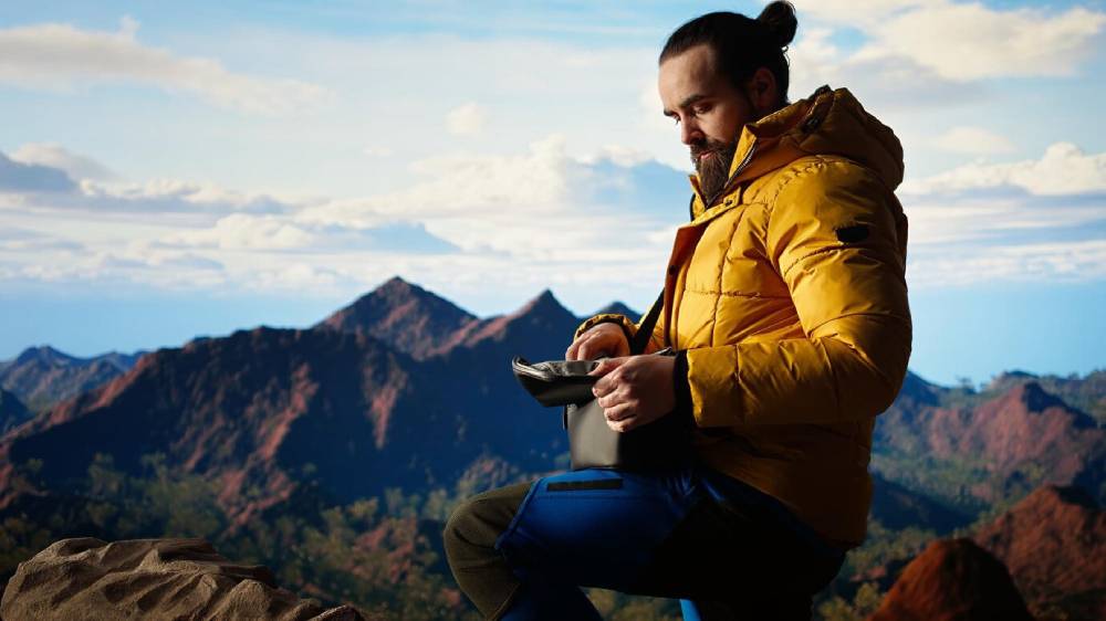 A person in a bright yellow jacket sits atop a rock, holding a device, with stunning mountains and a clear blue sky in the background.