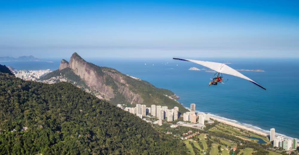 A hang glider soars above a coastal city, with green mountains and a prominent rock formation in the background under a clear blue sky.