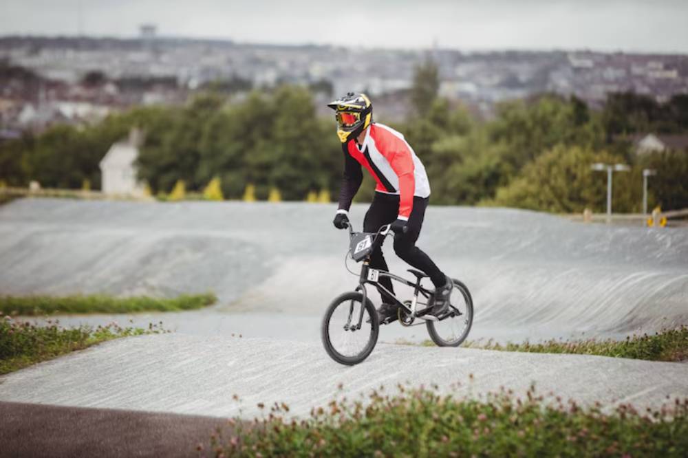 A BMX rider in protective gear performs a maneuver on a dirt track, surrounded by greenery and a cloudy skyline in the background.