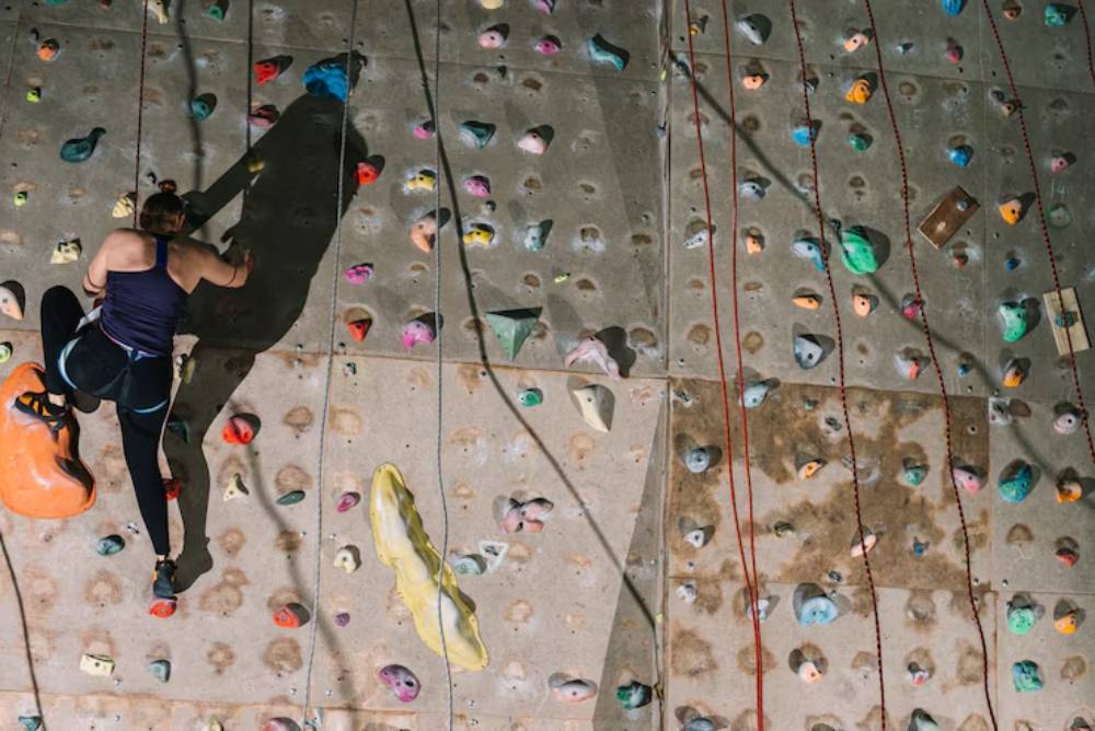A climber ascends a colorful indoor rock climbing wall, focused and determined, with gear and ropes visible in the background.