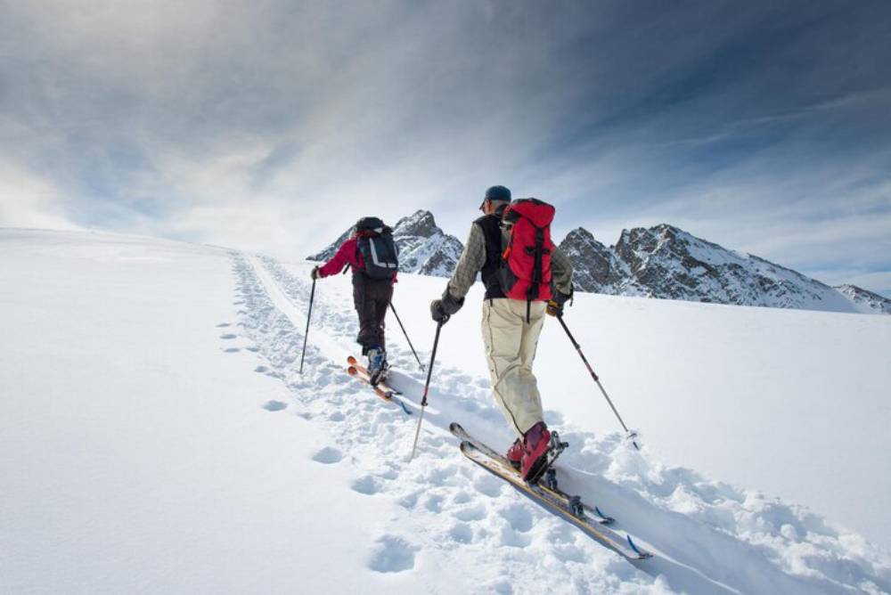 Two skiers ascend a snowy slope, leaving tracks behind them, with majestic mountains and a cloudy sky in the background.