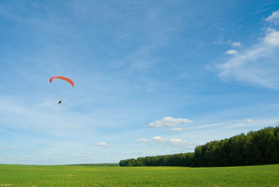 A paramotor with an orange wing soars over a lush green field under a bright blue sky dotted with clouds and a distant tree line.