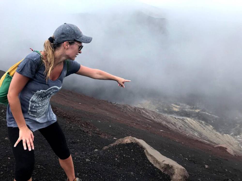 A woman in a gray t-shirt and cap points towards a smoky volcanic landscape shrouded in fog, showcasing her adventurous spirit.