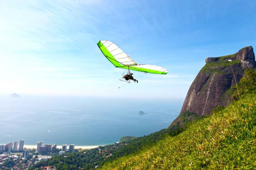 A hang glider soaring high above a coastal landscape, with lush green hills and a towering cliff in the background under a clear blue sky.