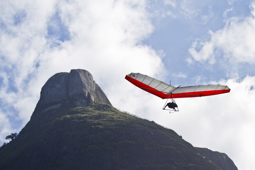 A hang glider with a red and white wing soars above a steep, rocky mountain under a partly cloudy sky.
