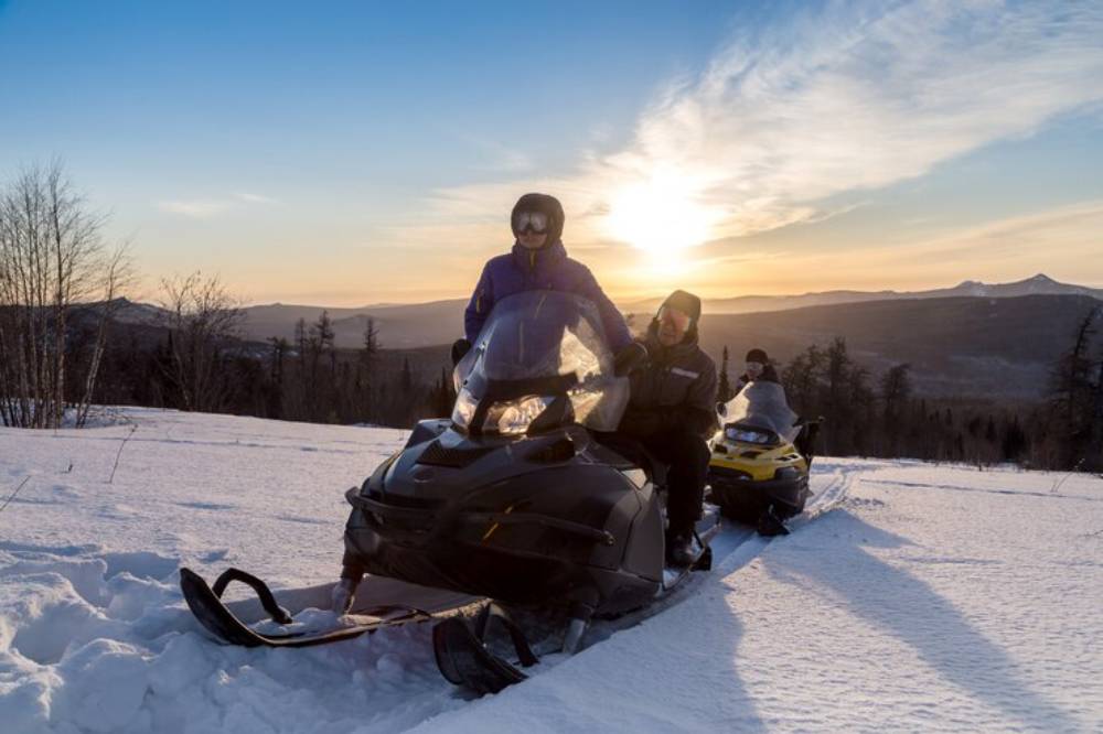 Two people on a snowmobile in a snowy landscape at sunset, surrounded by trees and mountains in the background.