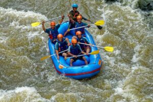 A group of six people in life vests and helmets navigate a blue raft through turbulent river rapids, paddles raised high.