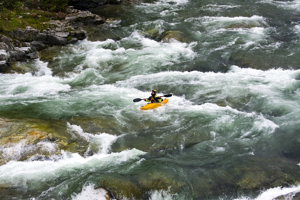 A kayaker navigates turbulent whitewater rapids in a bright yellow raft, surrounded by rocky banks and swirling green waters.
