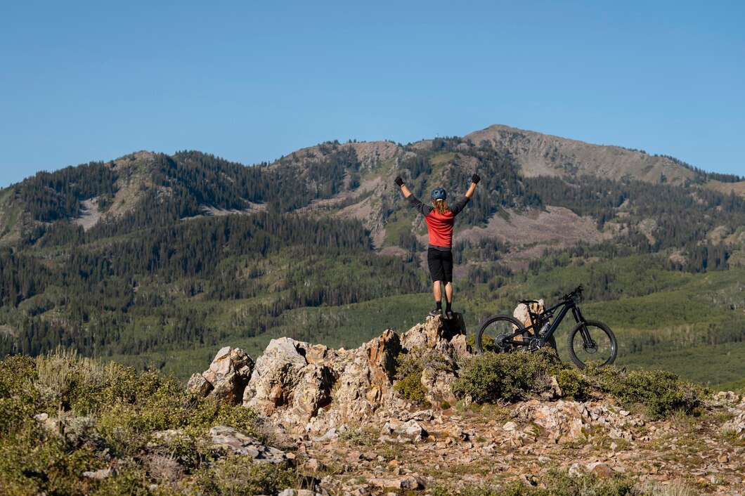A mountain biker stands triumphantly on a rock, arms raised, with a bike beside him and a lush mountainous landscape in the background.
