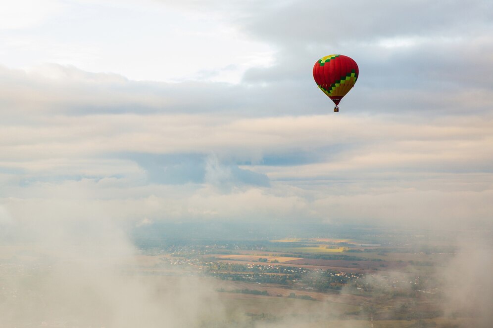 A colorful hot air balloon floats above a misty landscape, surrounded by soft clouds and a pale sky.