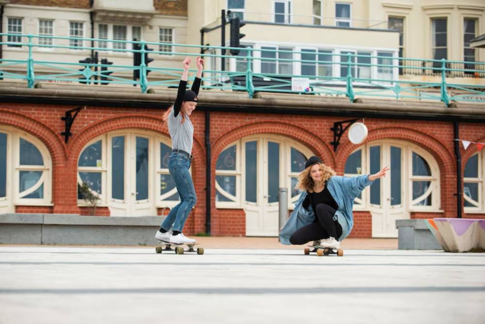 Two young women skateboarding on a sunny day in a vibrant urban setting, with stylish outfits and a playful atmosphere.