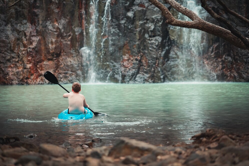 A boy paddles a bright blue kayak in tranquil water, with a scenic waterfall cascading down rocky cliffs in the background.