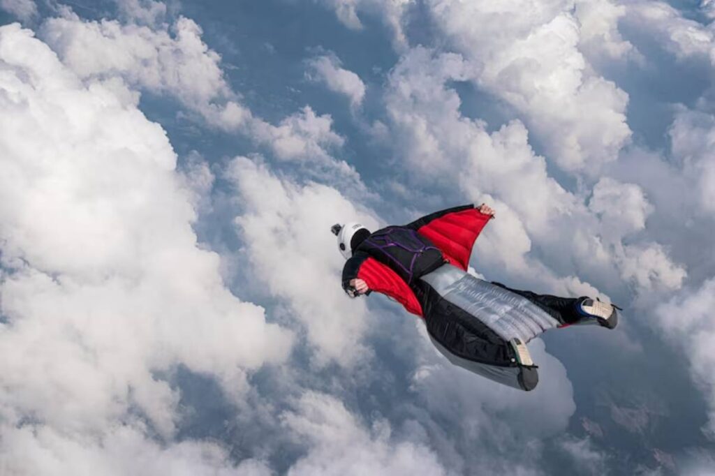 A wingsuit flyer glides effortlessly through fluffy clouds against a blue sky, showcasing the thrill of skydiving.