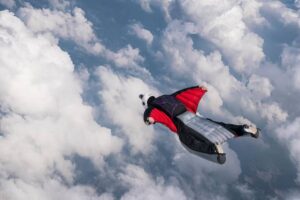A wingsuit flyer glides effortlessly through fluffy clouds against a blue sky, showcasing the thrill of skydiving.