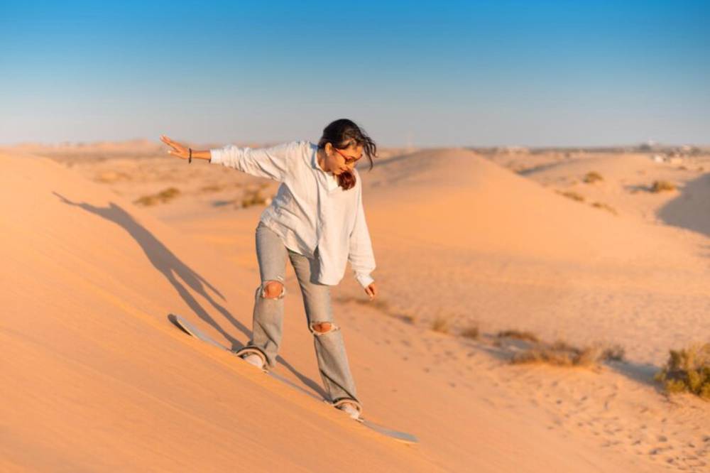 A person balances on a skateboard while sandboarding down a golden dune under a clear blue sky.
