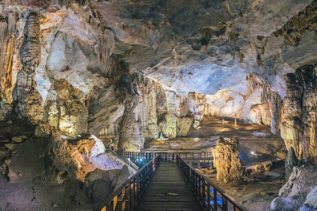 A scenic view of a cave featuring intricate stalactites and stalagmites, with a wooden walkway leading through the illuminated space.