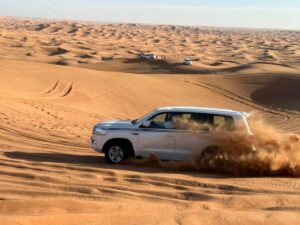 A white SUV driving through sandy dunes, kicking up dust, with distant vehicles visible in a vast desert landscape.