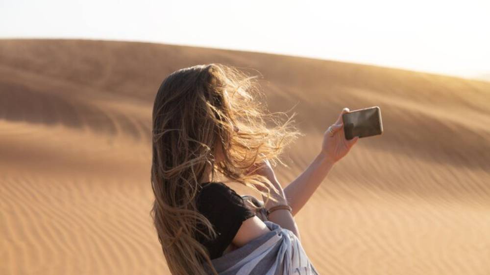 A woman with long hair captures a selfie in a sunlit desert, surrounded by rolling sand dunes.