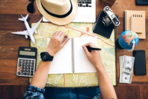 A person's hands jot down travel plans on a notebook, surrounded by a globe, calculator, camera, money, and a laptop on a map.