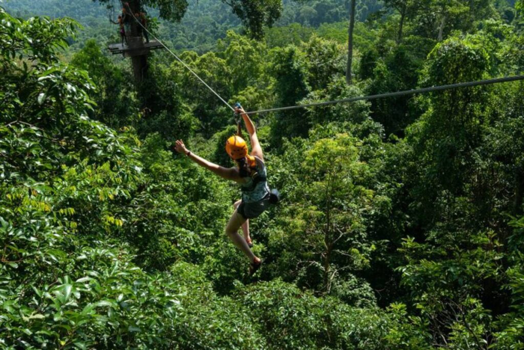 A thrill-seeker zip lines through lush green foliage, surrounded by dense jungle and distant hills, wearing a helmet and safety gear.