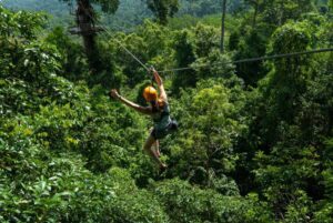 A thrill-seeker zip lines through lush green foliage, surrounded by dense jungle and distant hills, wearing a helmet and safety gear.